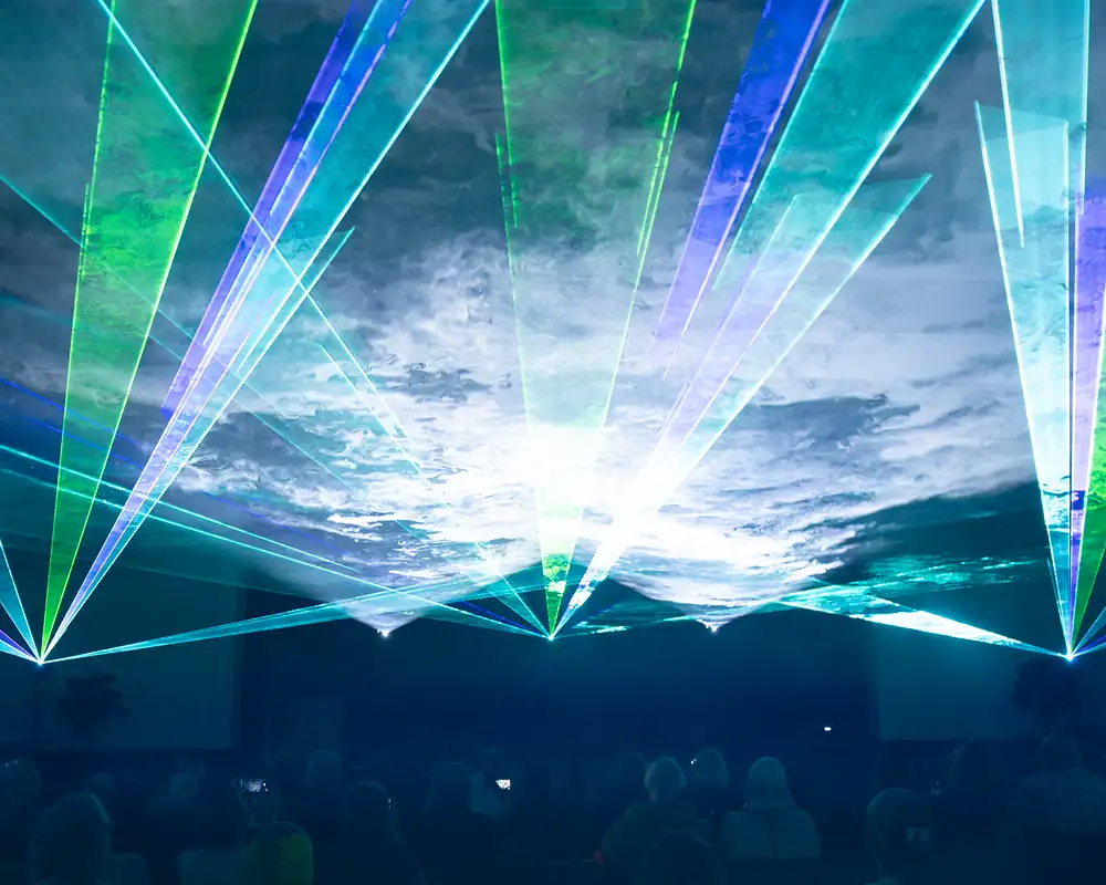 Cyanfarbene, grüne und blaue Laserfächer kreuzen sich über dem sitzenden Publikum, die Saaldecke leuchtet im Strahlenlicht