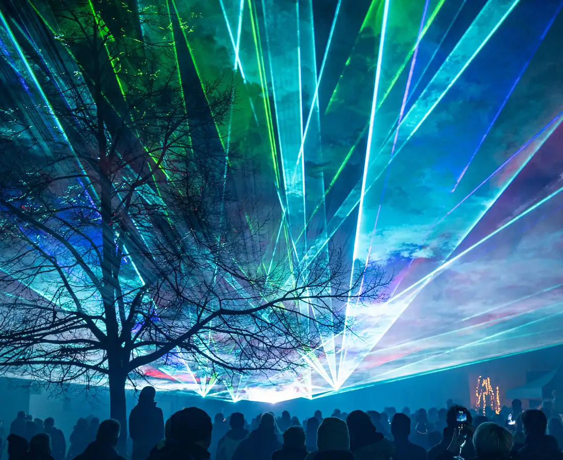 Mehrfarbiger Laserfächer in Cyan, Blau, Grün und Magenta füllt den Himmel über dem Silvesterpublikum und dem Dorfbaum