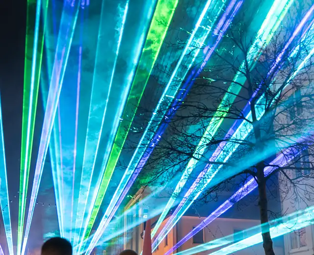Fächer aus scharfen cyan-, blau- und grünfarbenen Laserlinien über der Singener Fußgängerzone, dazwischen ein kahler Baum